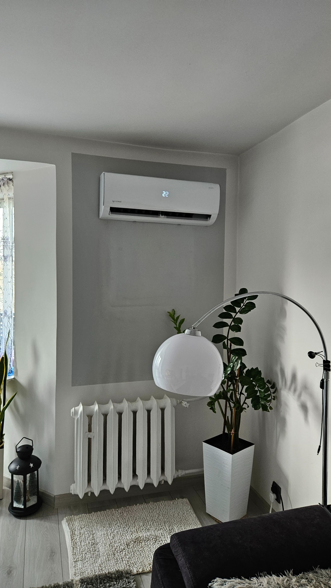 Biały klimatyzator ścienny z wyświetlaczem temperatury, zamontowany na szarej ścianie w salonie w Górze, widoczny obok grzejnika i lampy podłogowej, z rośliną doniczkową w tle.