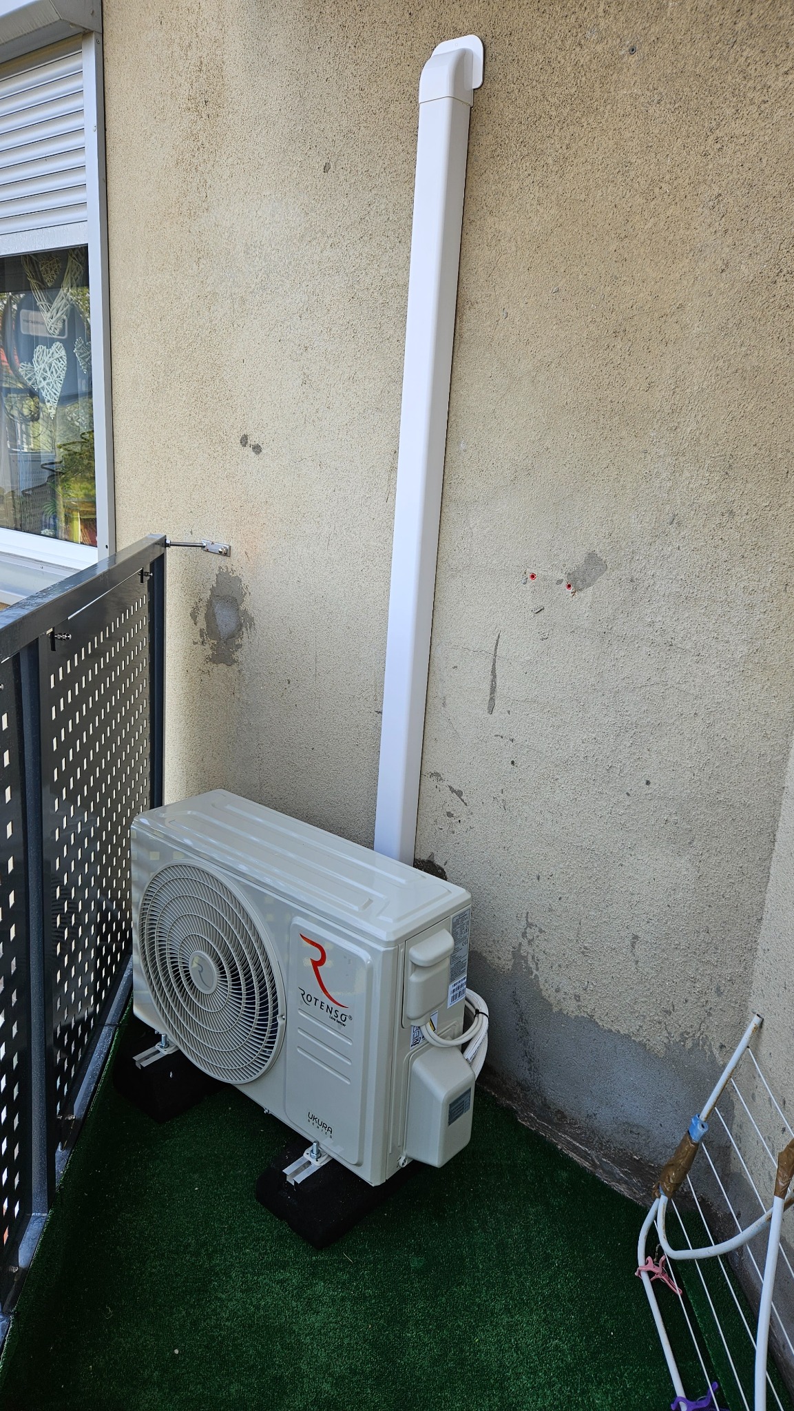 Zewnętrzna jednostka klimatyzacji Rotenso Ukura zamontowana na balkonie z imitacją trawy, widoczna biała rura odprowadzająca skropliny oraz ślady po wcześniejszych mocowaniach na ścianie.