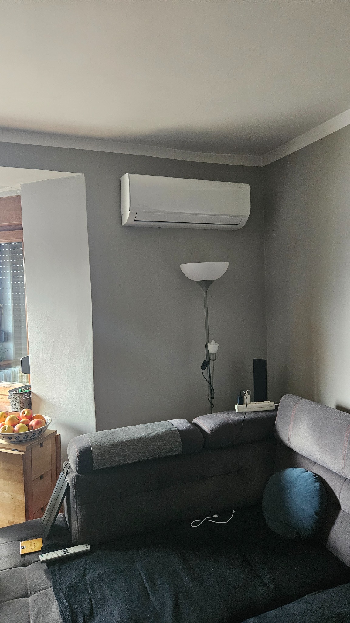 Biała jednostka klimatyzacyjna zamontowana na szarej ścianie w pokoju, widoczna nad szarą sofą z poduszką i lampą stojącą, z owocami w misce w tle.