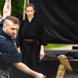 Mężczyzna w stroju do ćwiczeń przecina mieczem bambusową matę, drugi mężczyzna w tle obserwuje, widoczny tatuaż na ramieniu.