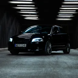 Czarne auto marki Audi A3 stoi w ciemnym garażu oświetlonym rzędami lamp jarzeniowych, widoczne światła przednie i tablica rejestracyjna WE 1J248.