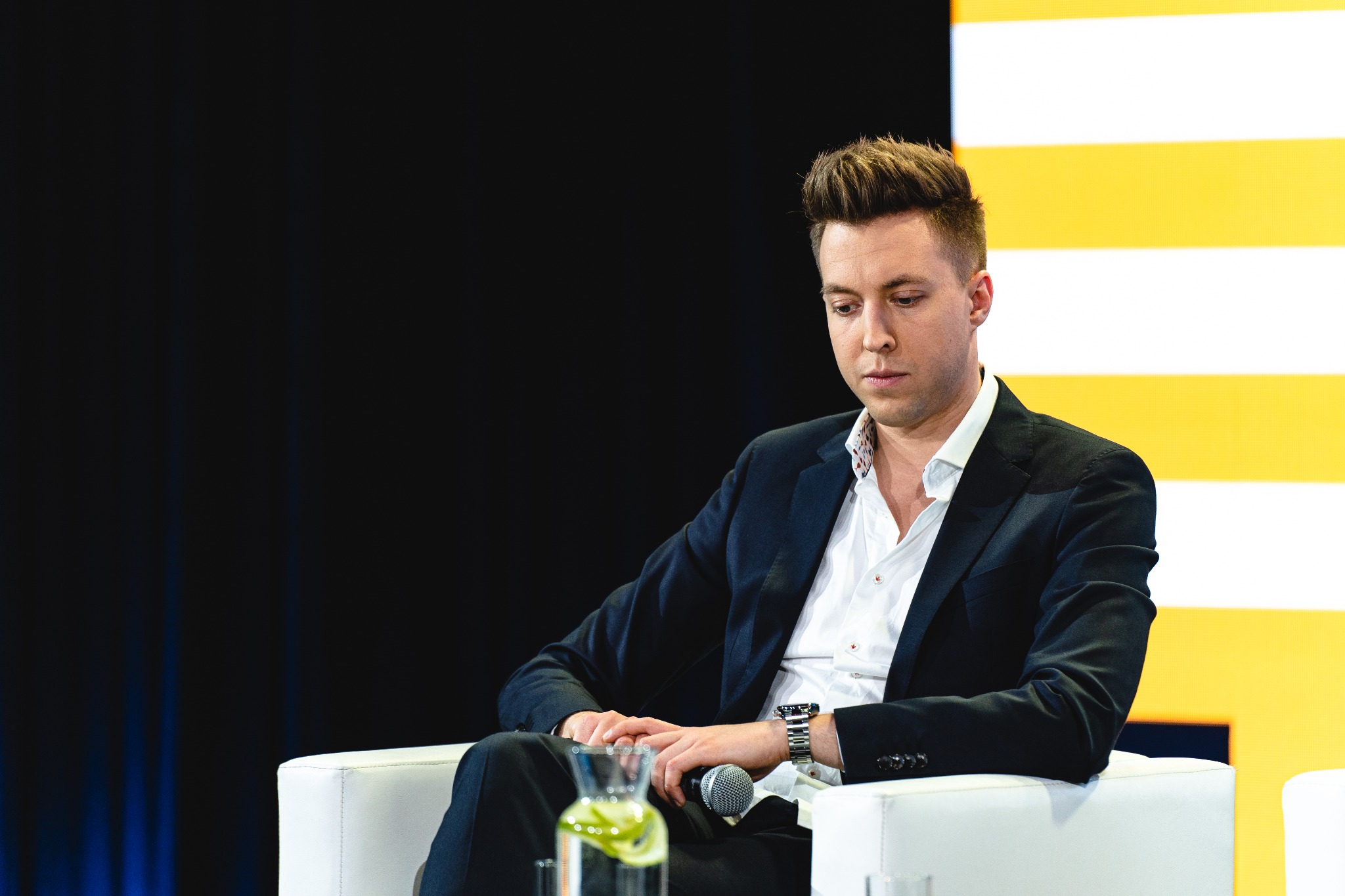 Mężczyzna w garniturze siedzi na białym krześle, trzyma mikrofon, w tle czarno-żółte pasy, na stole dzbanek z wodą i cytryną.