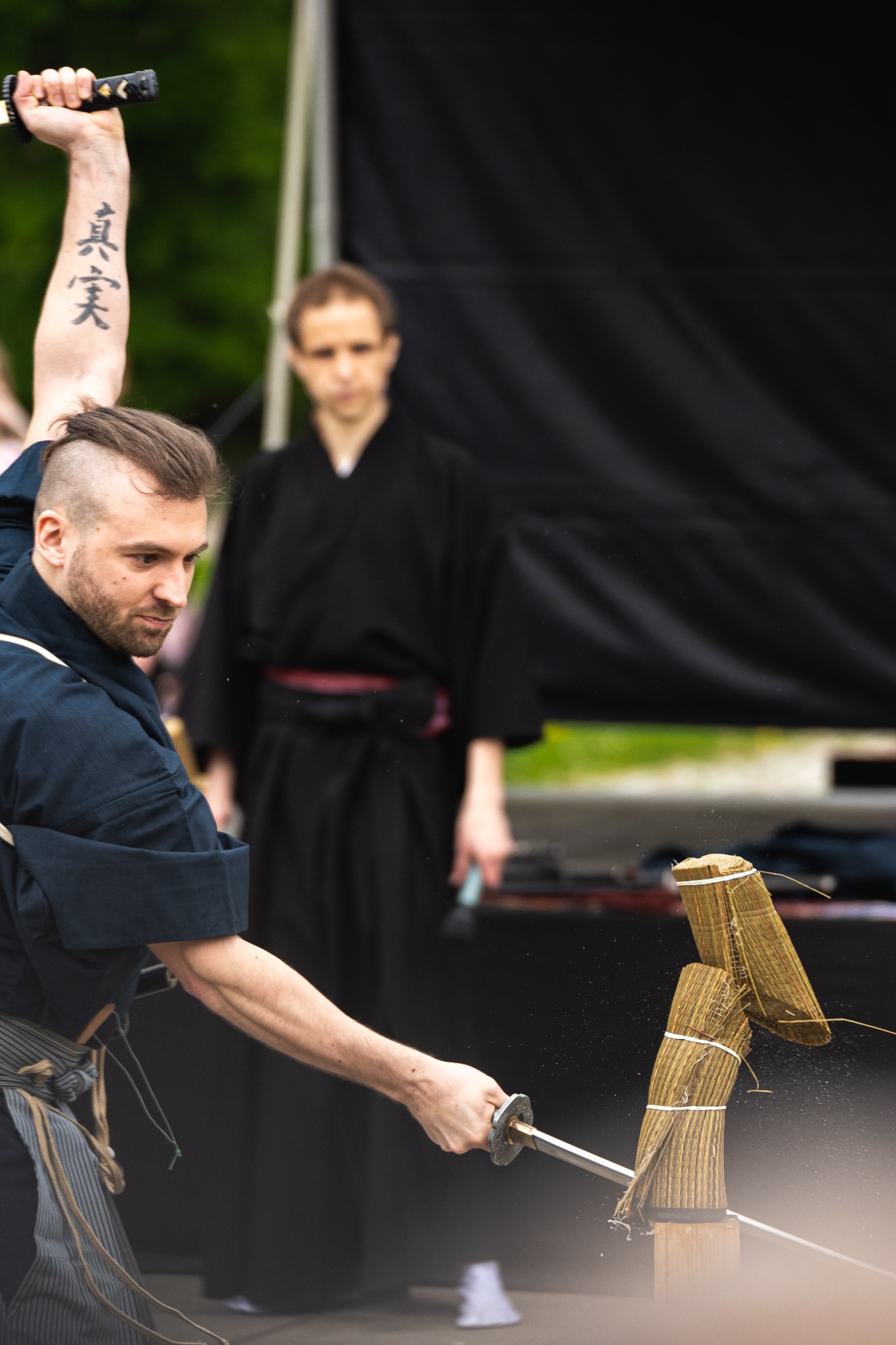 Mężczyzna w stroju do ćwiczeń przecina mieczem bambusową matę, drugi mężczyzna w tle obserwuje, widoczny tatuaż na ramieniu.