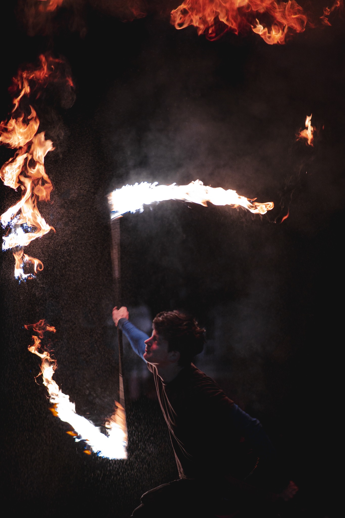 Młody mężczyzna w ciemnym otoczeniu wykonuje pokaz z płonącymi pochodniami, otoczony iskrami i płomieniami, z twarzą skierowaną ku górze.