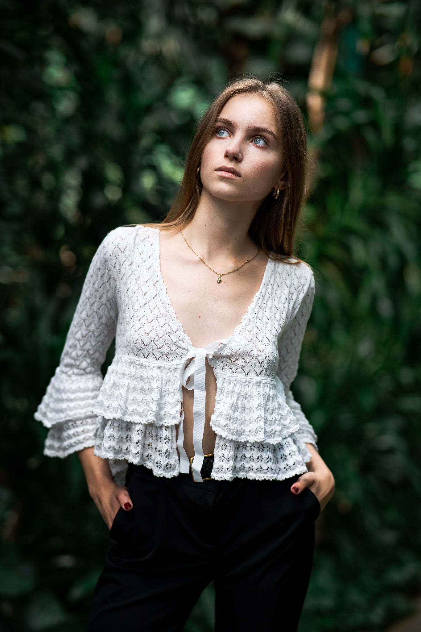 Portret młodej kobiety z jasnymi oczami, ubrana w koronkową bluzkę i czarne spodnie, pozująca na tle zieleni, z rękami w kieszeniach i wzrokiem skierowanym w górę.