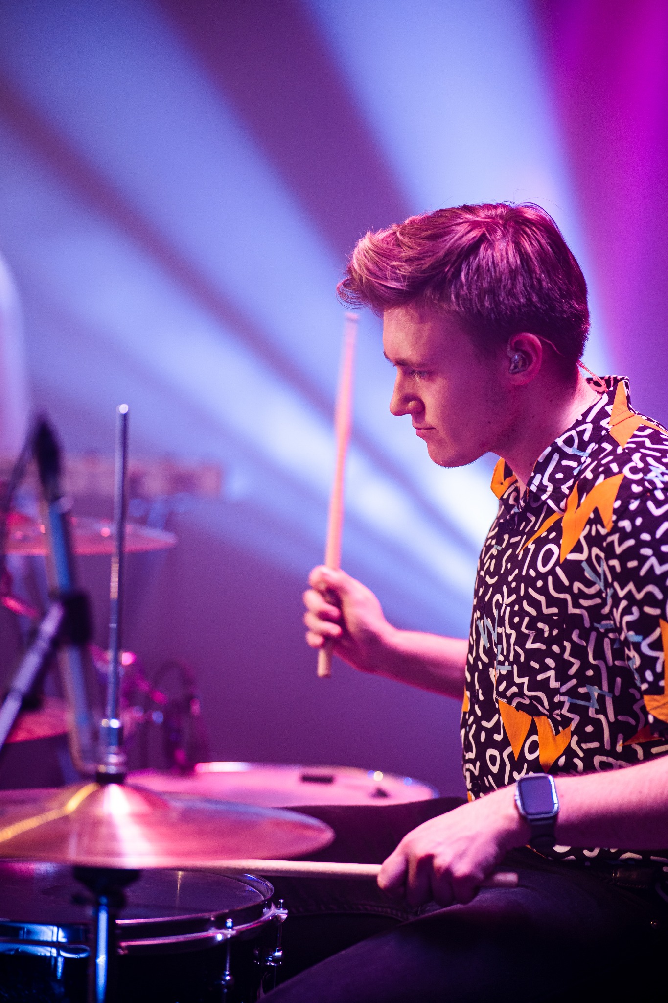 Młody perkusista w koszuli we wzory uderza pałeczką w talerz perkusyjny pod fioletowo-niebieskim oświetleniem scenicznym.