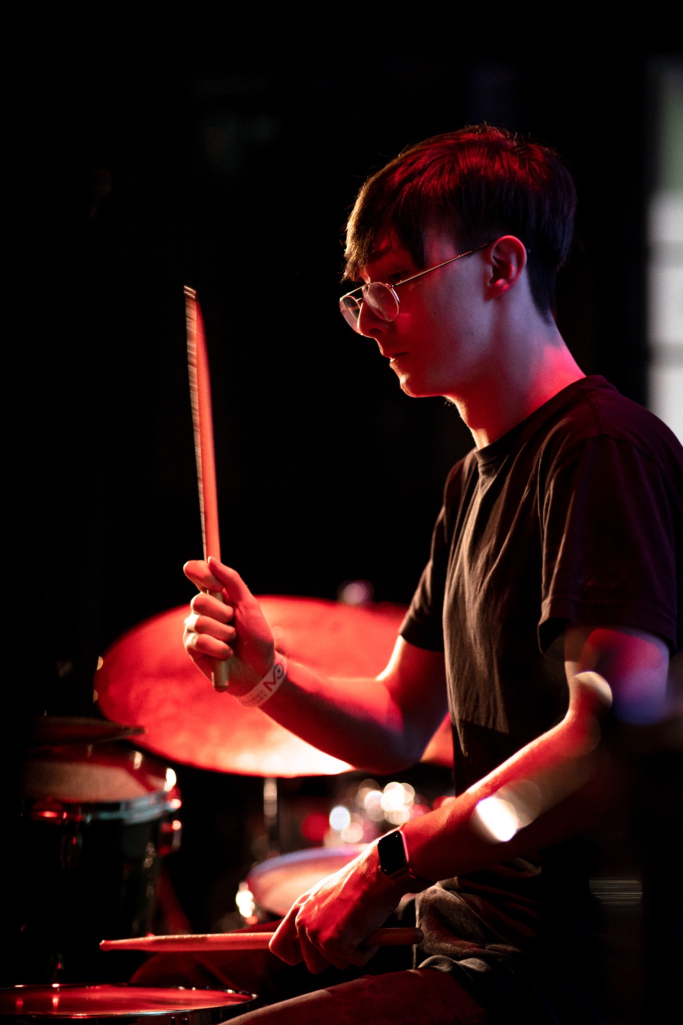 Młody perkusista w okularach, grający na perkusji w czerwonym oświetleniu scenicznym w Warszawie. Widoczny detal zegarka na nadgarstku.