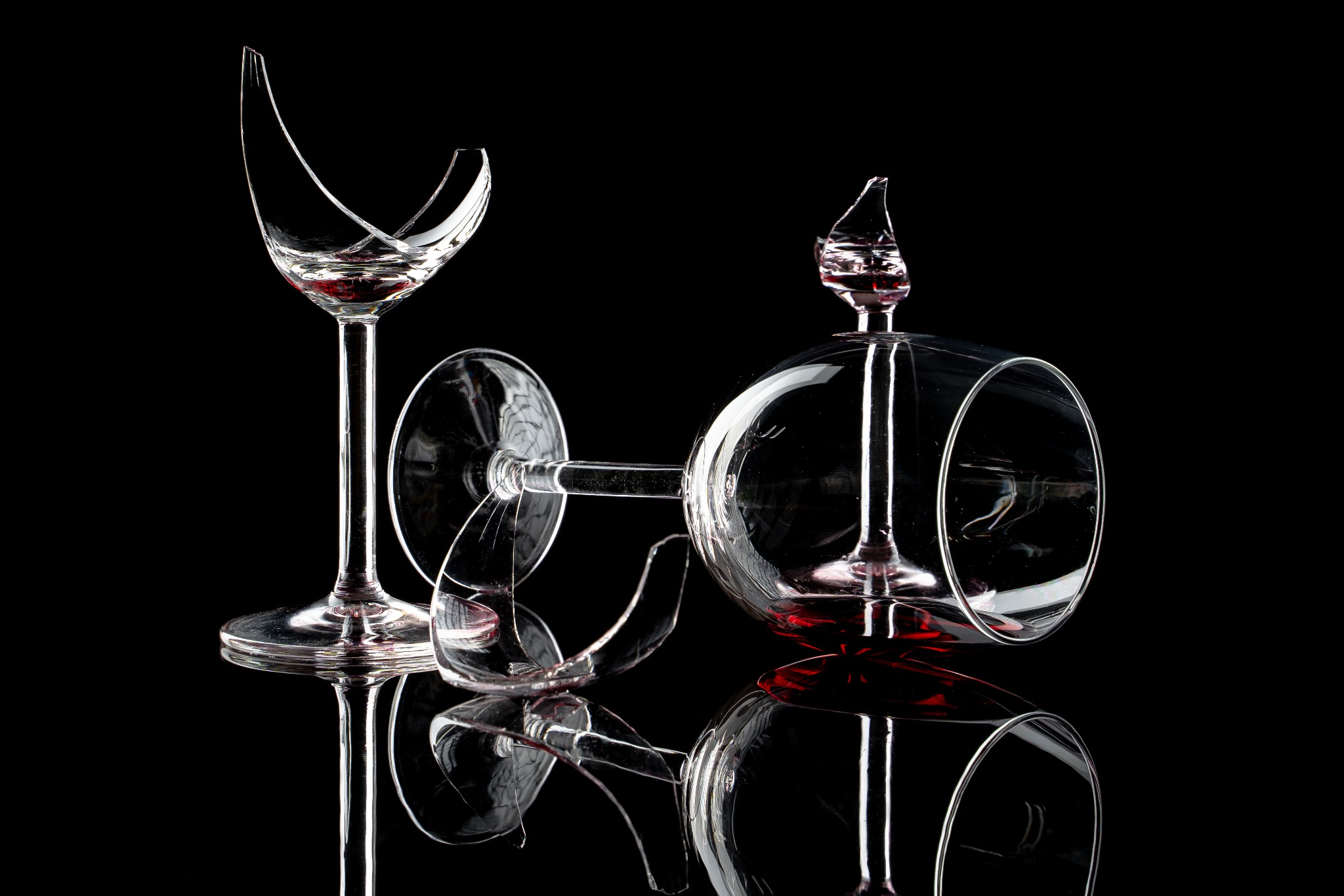 Dwa potłuczone kieliszki do wina na czarnym, błyszczącym tle, z widocznymi drobnymi odłamkami szkła i resztkami czerwonego wina.