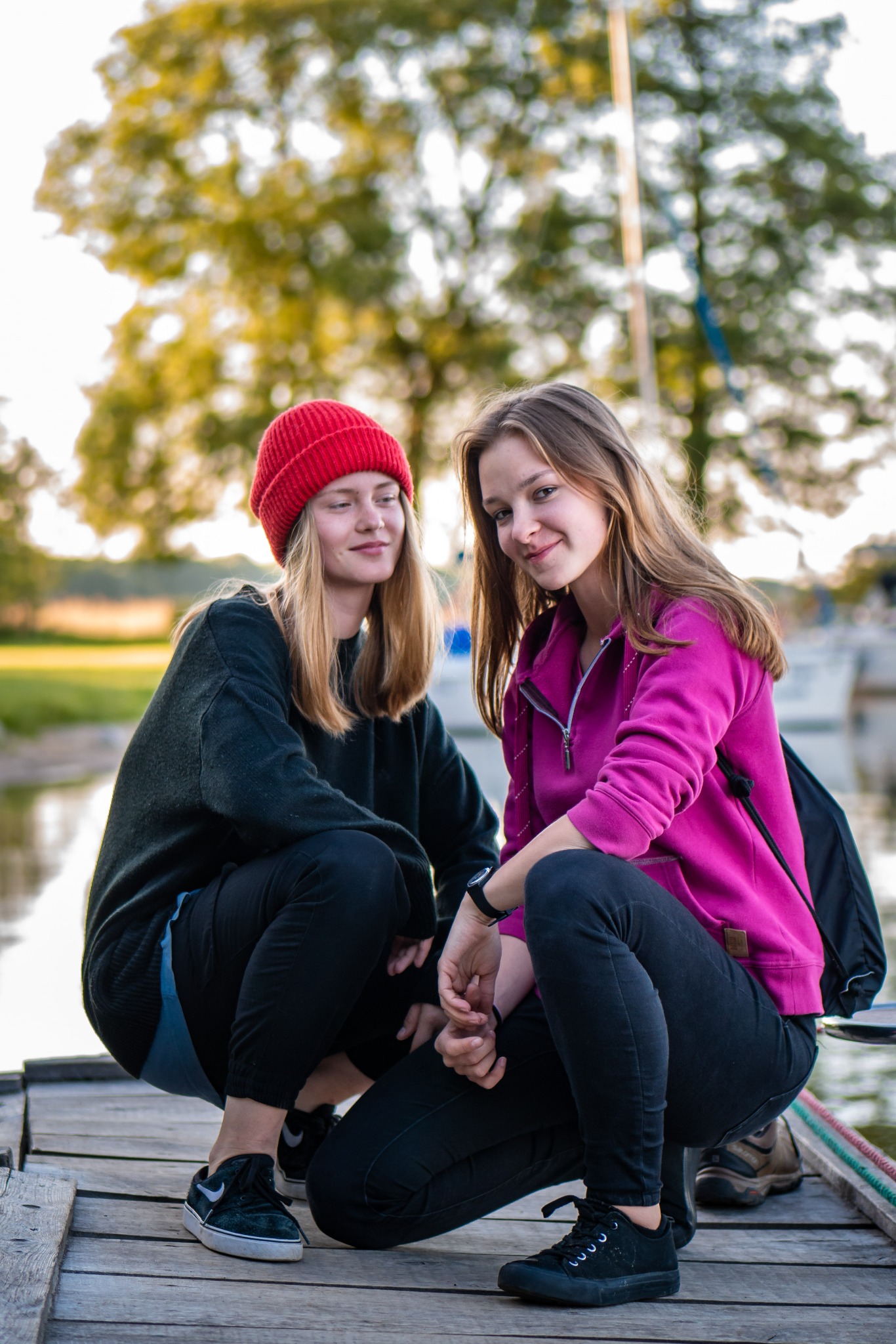 Dwie młode kobiety w kucającej pozycji na drewnianym pomoście, w tle woda i jacht, jedna w czerwonej czapce i czarnej bluzie, druga w różowej bluzie, obie uśmiechnięte.