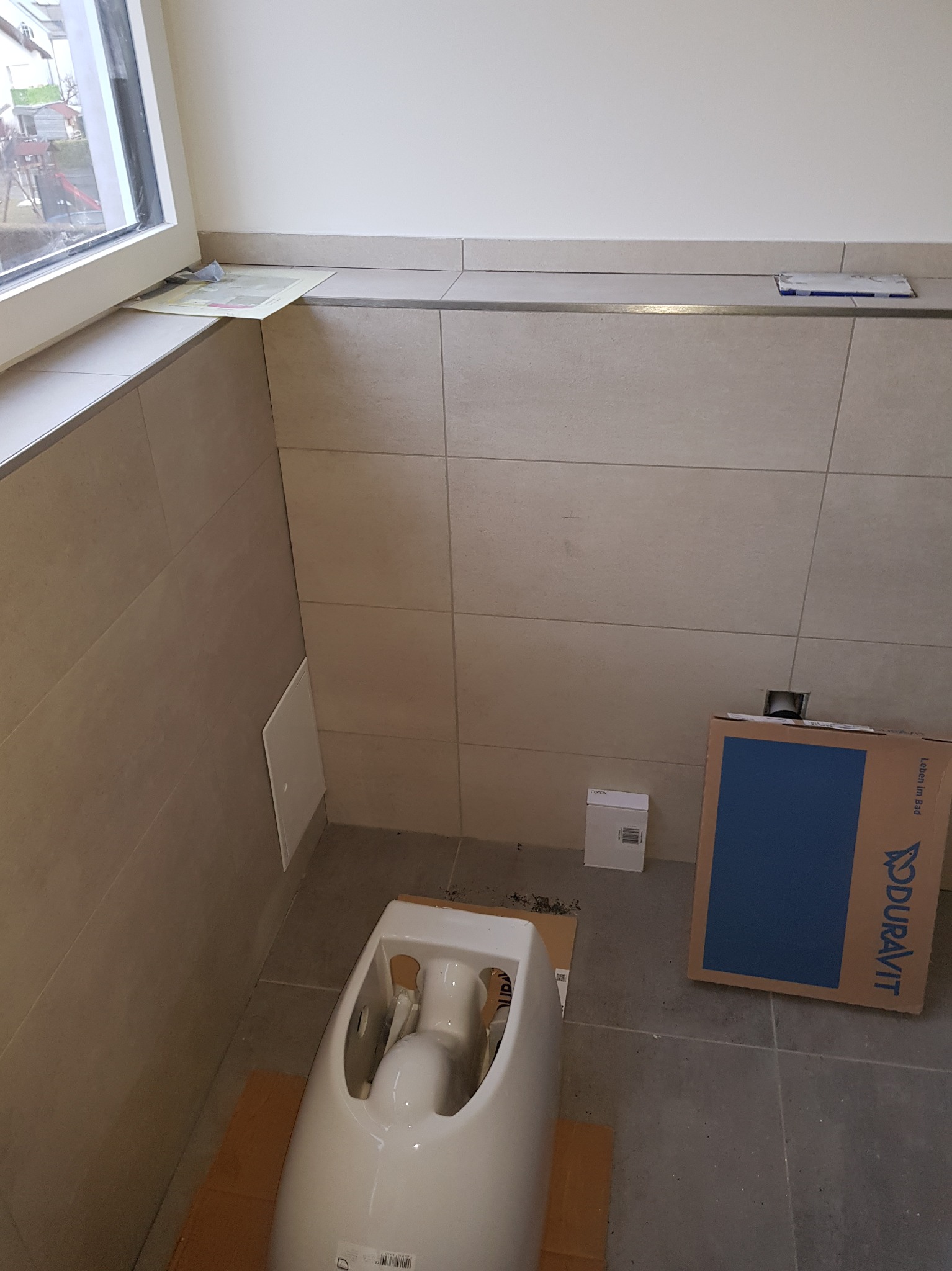 Montaż podwieszanej miski WC Duravit w łazience z beżowymi płytkami, widoczny karton i akcesoria montażowe.