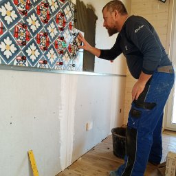 Sławomir Fornal TASMAN - Mężczyzna w trakcie układania dekoracyjnych płytek ściennych z użyciem systemu poziomowania, widoczne krzyżyki dystansowe i metalowy profil montażowy.