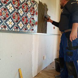 Sławomir Fornal TASMAN - Mężczyzna w kombinezonie nakłada klej na ścianę pod płytki dekoracyjne z wzorem kwiatowym, używając szpachli z zębami i poziomicy laserowej.