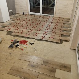 Sławomir Fornal TASMAN - Układanie drewnopodobnych płytek podłogowych z użyciem systemu poziomowania, widoczne narzędzia i akcesoria glazurnicze, surowe ściany i podłoga w tle.