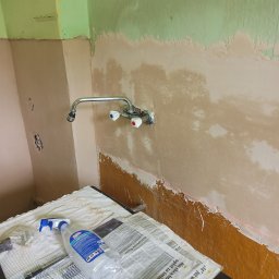 Sławomir Fornal TASMAN - Przygotowanie ściany do prac wykończeniowych w Marciszowie: widoczne ślady po szpachlowaniu, fragment starej farby, laserowy poziom, bateria umywalkowa i narzędzia na podłodze zabezpieczonej gazetami.