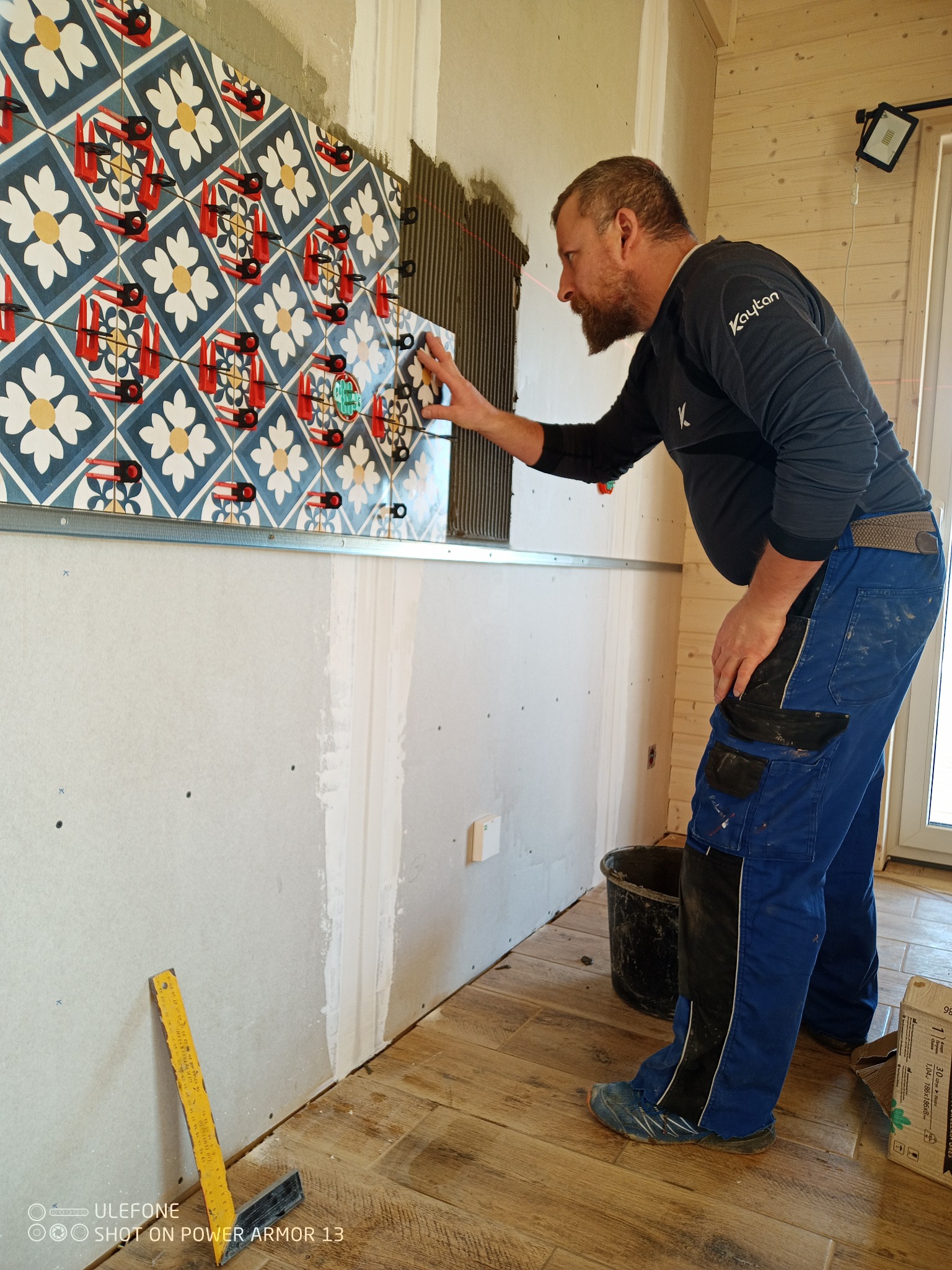 Mężczyzna w trakcie układania dekoracyjnych płytek ściennych z użyciem systemu poziomowania, widoczne krzyżyki dystansowe i metalowy profil montażowy.