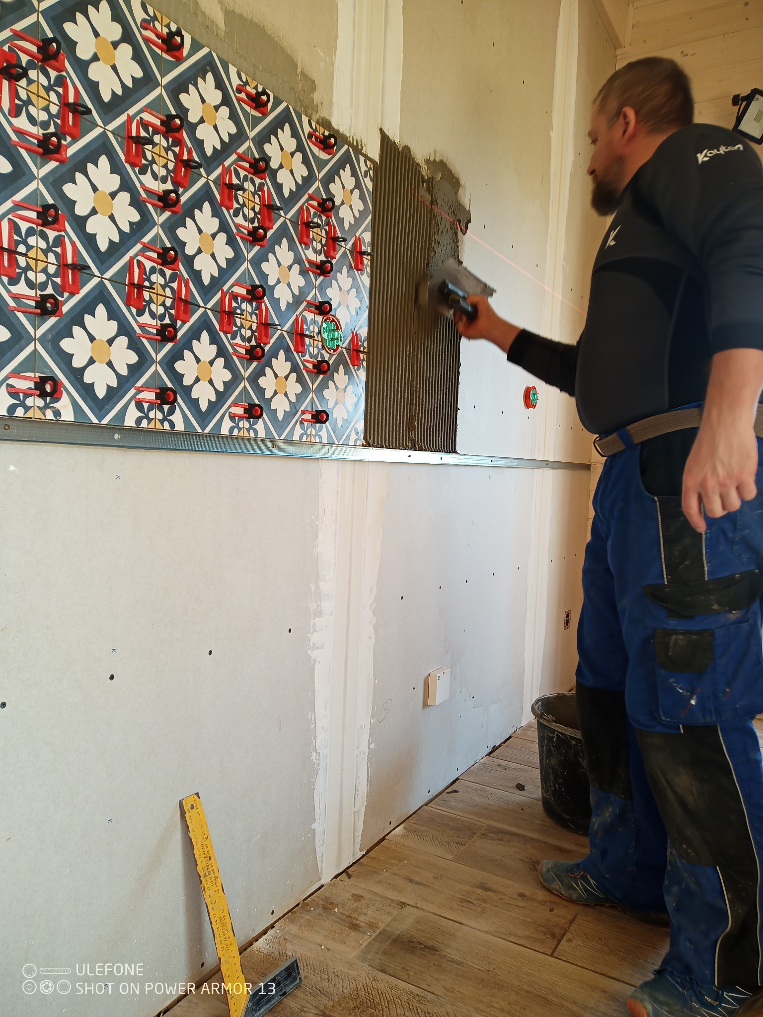 Mężczyzna w kombinezonie nakłada klej na ścianę pod płytki dekoracyjne z wzorem kwiatowym, używając szpachli z zębami i poziomicy laserowej.