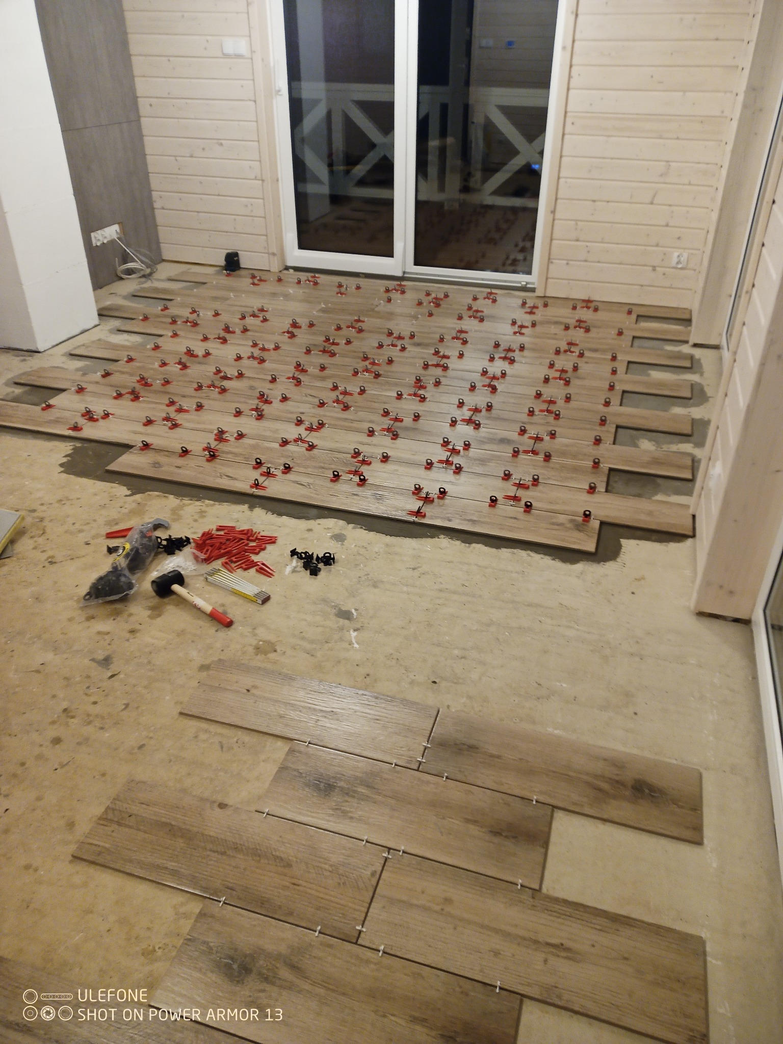 Układanie drewnopodobnych płytek podłogowych z użyciem systemu poziomowania, widoczne narzędzia i akcesoria glazurnicze, surowe ściany i podłoga w tle.