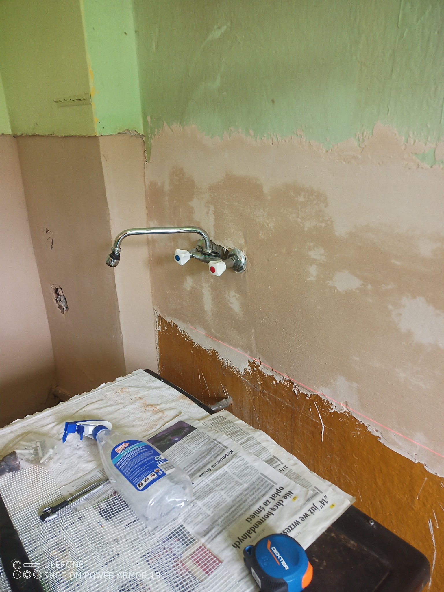 Przygotowanie ściany do prac wykończeniowych w Marciszowie: widoczne ślady po szpachlowaniu, fragment starej farby, laserowy poziom, bateria umywalkowa i narzędzia na podłodze zabezpieczonej gazetami.