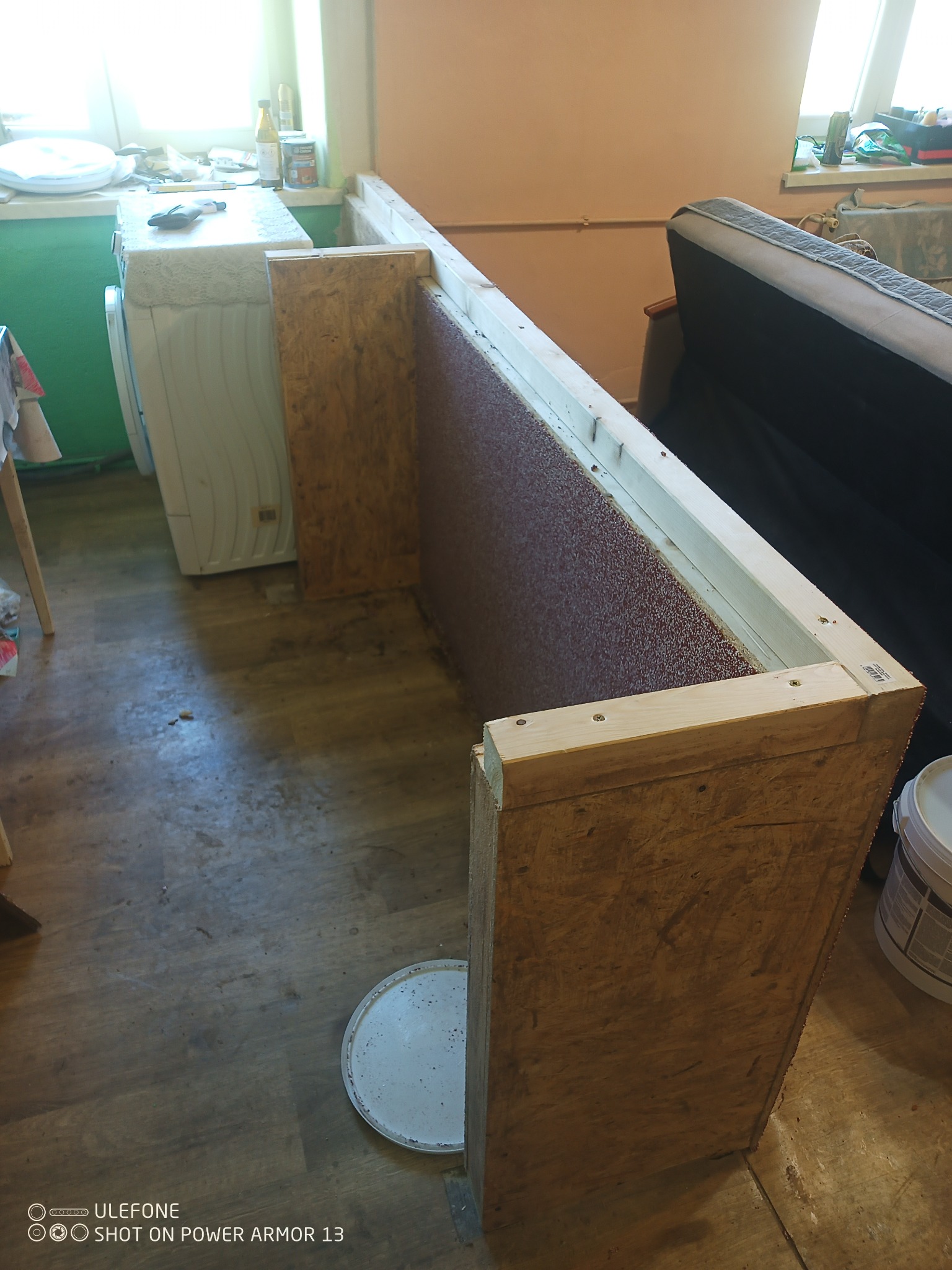 Konstrukcja z płyty OSB i drewnianych belek w trakcie budowy, prawdopodobnie obudowa w łazience, widoczna pralka i fragment sofy w tle.