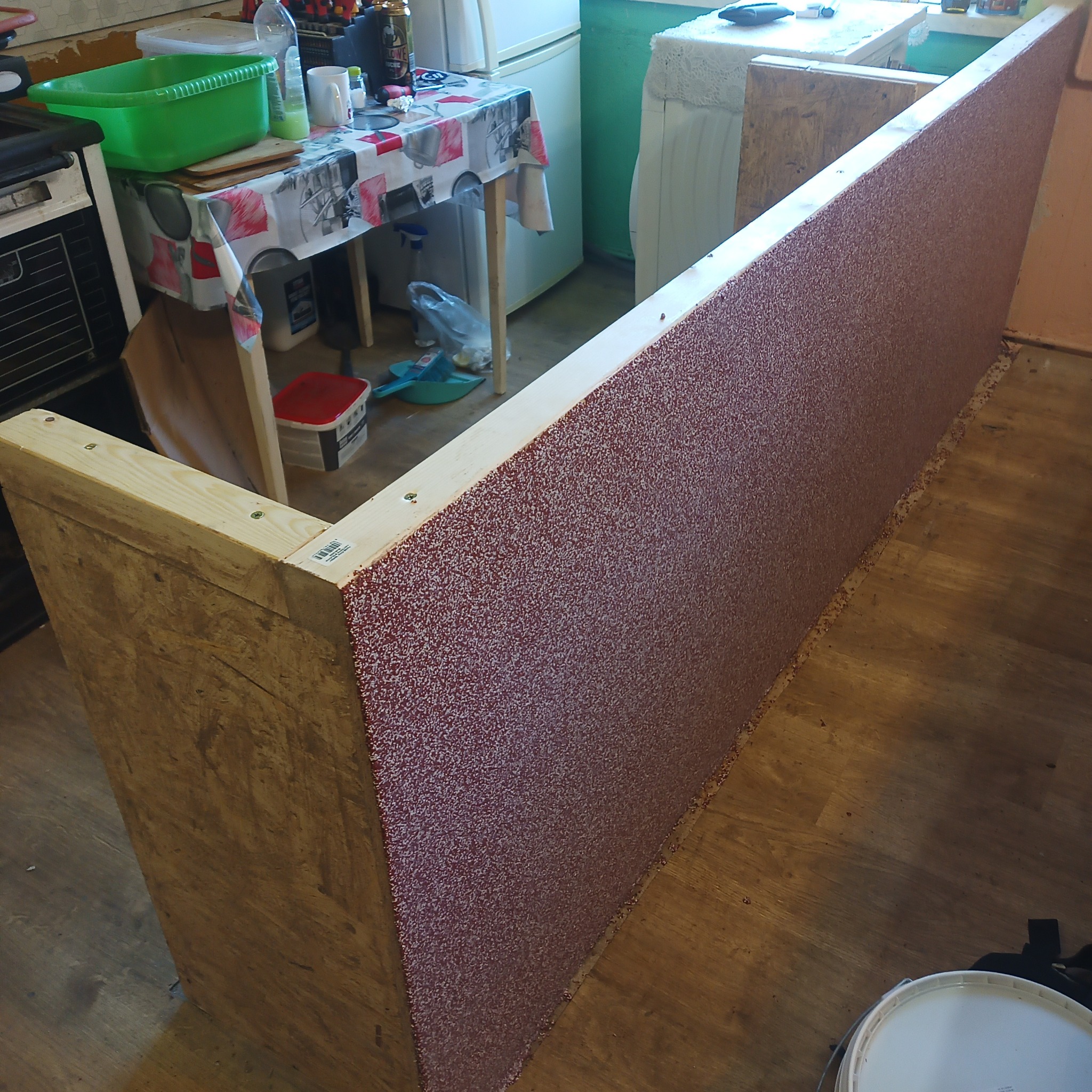 Konstrukcja z drewnianych belek i płyty OSB pokrytej czerwoną, ziarnistą strukturą, prawdopodobnie przygotowana do dalszych prac wykończeniowych w pomieszczeniu z linoleum na podłodze.