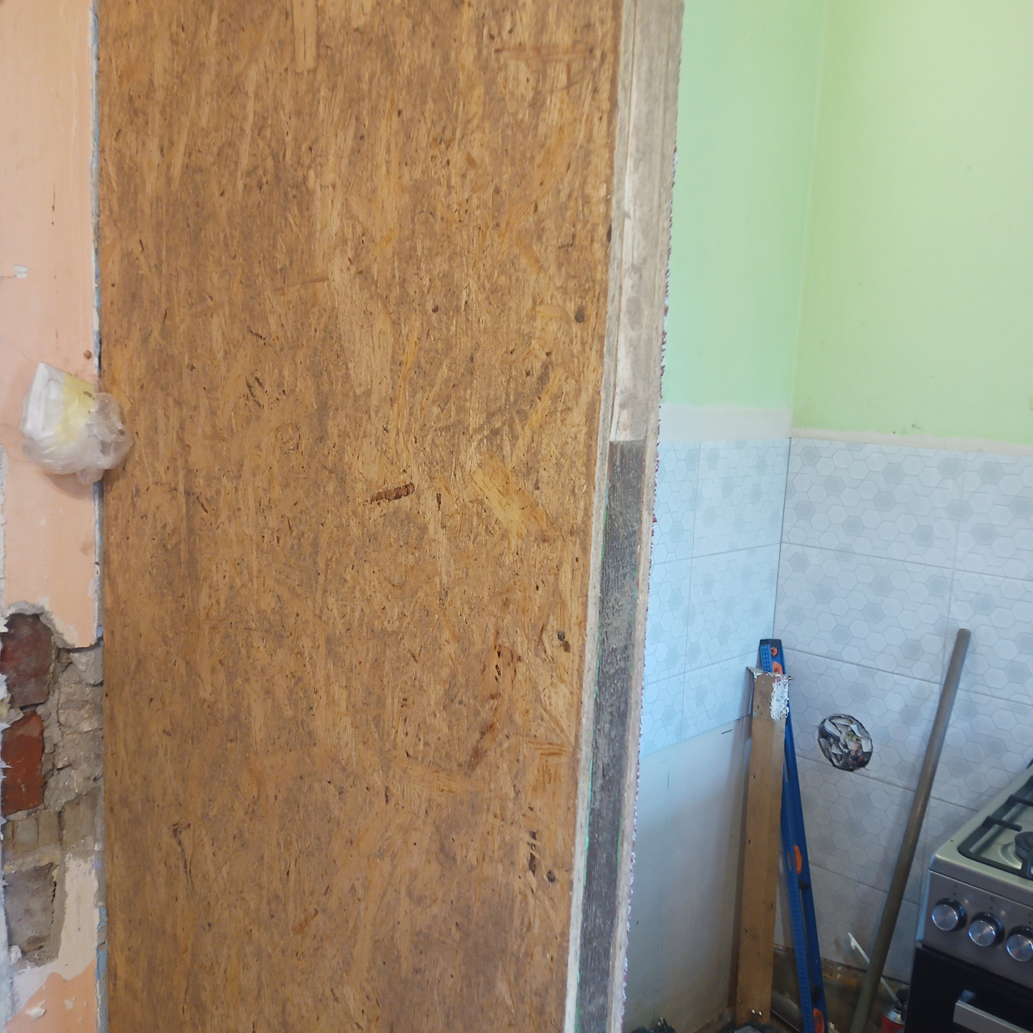 Płyta OSB zasłaniająca fragment ściany w trakcie prac remontowych w kuchni, widoczny fragment ściany z cegły, poziomica, fragment kuchenki gazowej i białe heksagonalne płytki na ścianie.