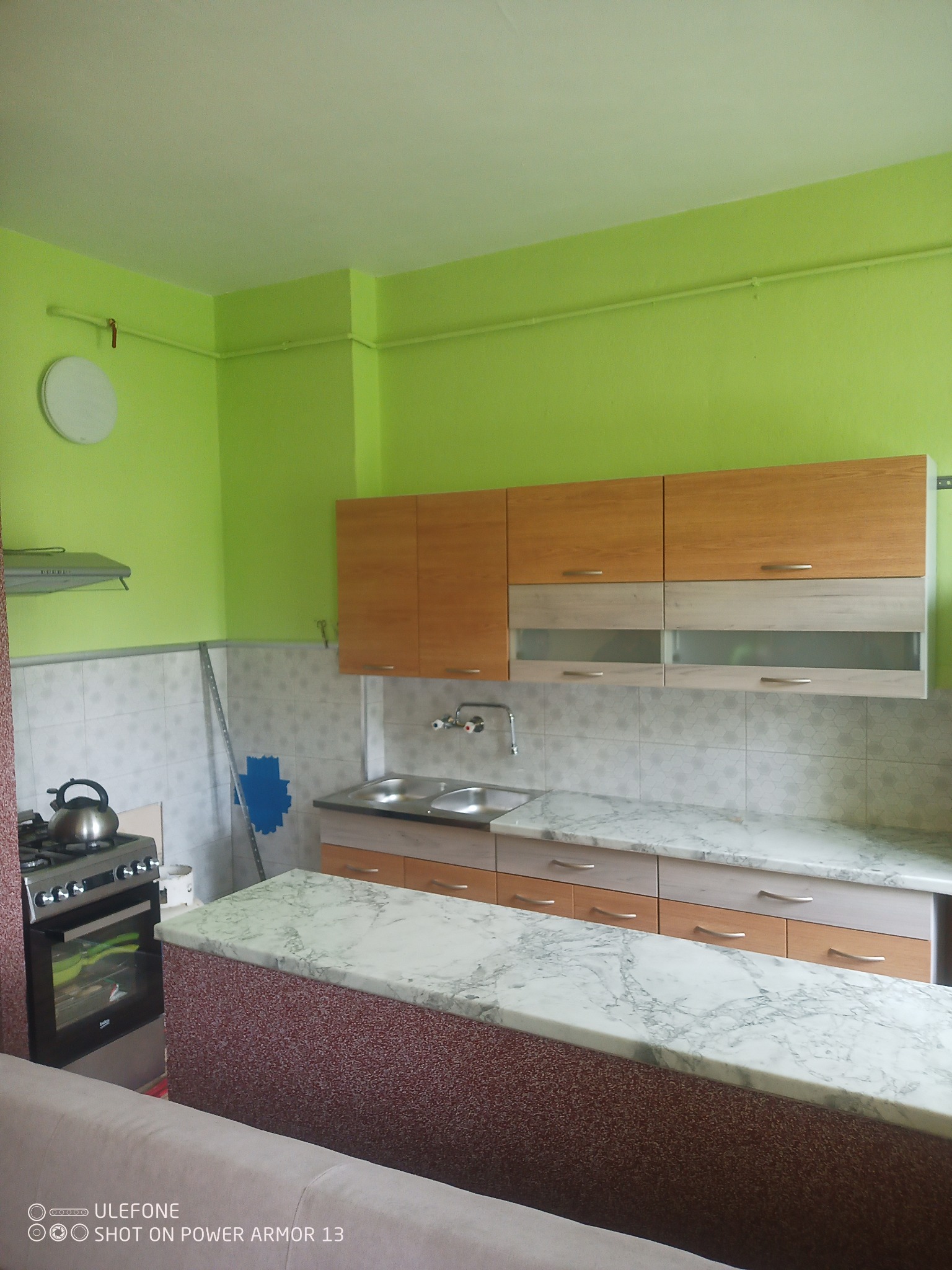 Fragment kuchni z zielonymi ścianami, jasnymi szafkami i blatem imitującym marmur, widoczny piec z czajnikiem i fragment sofy w dolnej części kadru.