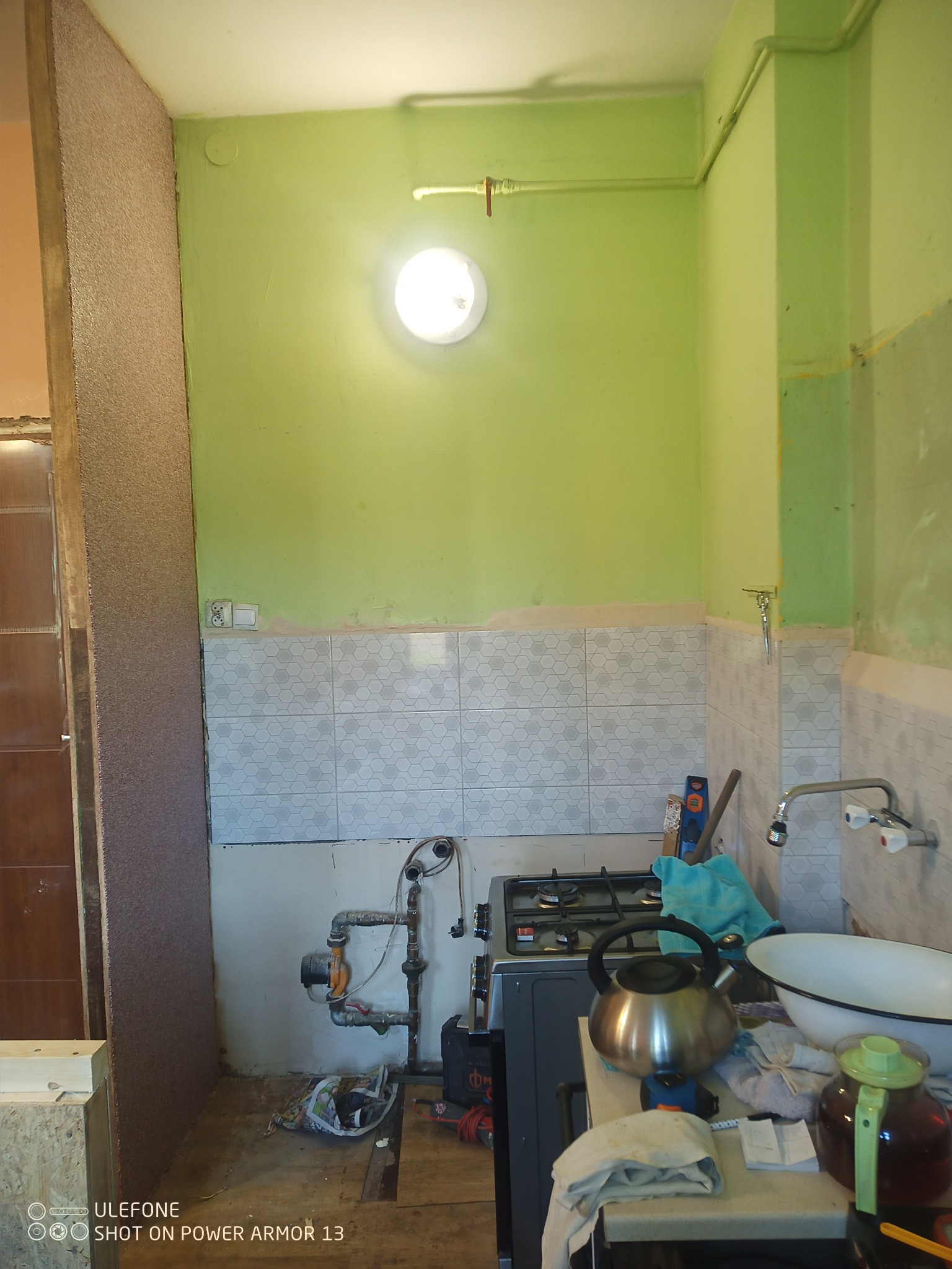 Wnętrze kuchni w trakcie remontu: częściowo położone białe płytki heksagonalne, odsłonięte rury gazowe, kuchenka gazowa i prowizoryczne wyposażenie na blacie.