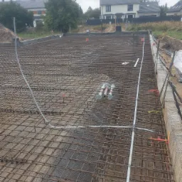 Zbrojenie fundamentów z widocznymi rurami instalacji elektrycznej, przygotowane do zalania betonem, na tle budynków mieszkalnych.