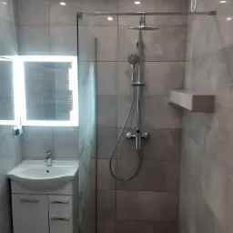 Nowoczesna łazienka z szarymi płytkami, kabiną prysznicową ze szklaną ścianką, podświetlanym lustrem nad białą szafką z umywalką i półką pod prysznicem.