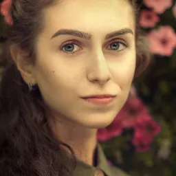 Portret młodej kobiety o niebieskich oczach, z ciemnymi włosami upiętymi z tyłu, na tle różowych kwiatów i zieleni, ubrana w oliwkową koszulę z drewnianymi guzikami.