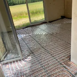 Białe rurki ogrzewania podłogowego ułożone na srebrnej folii z czerwoną kratką w surowym wnętrzu z widokiem na pole przez duże okno.