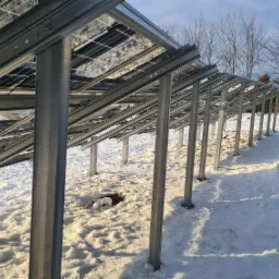 Rząd paneli fotowoltaicznych zamontowanych na metalowej konstrukcji w zimowym krajobrazie, śnieg wokół podstaw słupków, w tle drzewa i gabionowy mur.