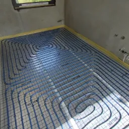 Niebieskie rury ogrzewania podłogowego marki Keller ułożone na izolacji z siatką, widok z góry na surowy beton i okno