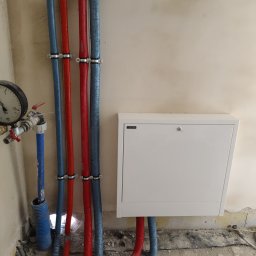 Hydrokwiat Robert Kwiatkowski - Węzeł instalacyjny z manometrem, rurami w kolorach niebieskim i czerwonym oraz białą szafką rewizyjną na tle surowej ściany.
