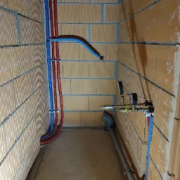 Instalacja wodno-kanalizacyjna i grzewcza w surowym pomieszczeniu we Wrocławiu, widoczne rury w izolacji termicznej w kolorach czerwonym i niebieskim, manometry oraz odpływ kanalizacyjny.