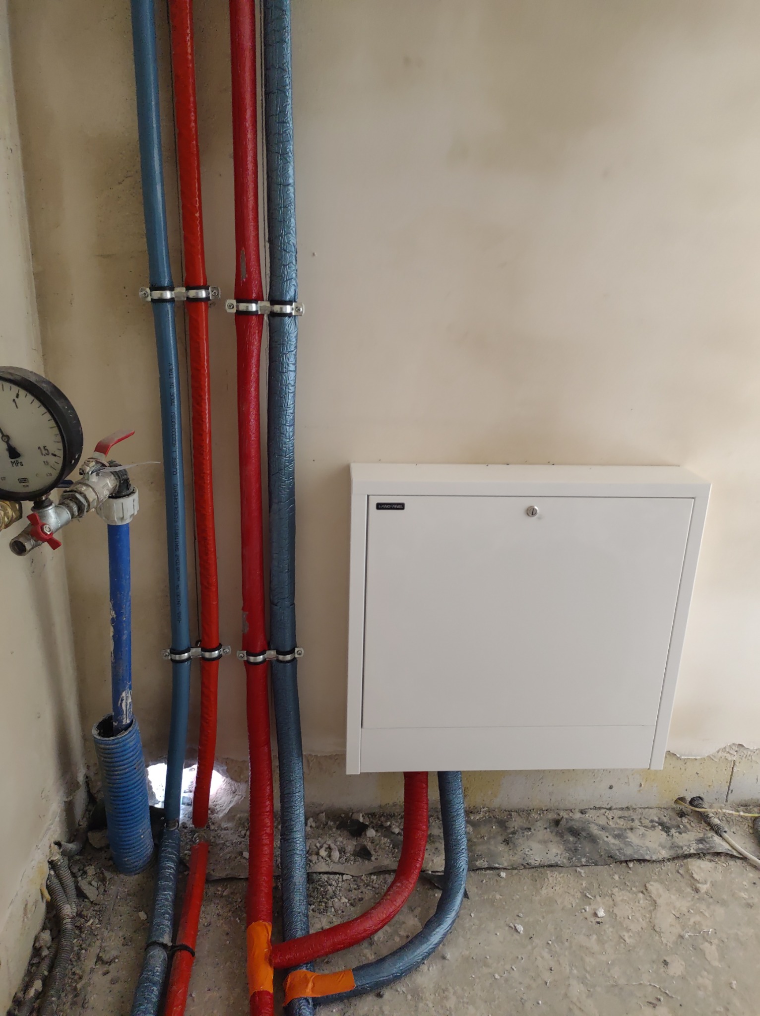 Węzeł instalacyjny z manometrem, rurami w kolorach niebieskim i czerwonym oraz białą szafką rewizyjną na tle surowej ściany.