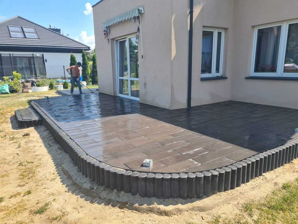 Umyta, brązowa kostka brukowa ułożona na tarasie, z obrzeżem z okrąglaków, pracownik w tle zamiata wodę.