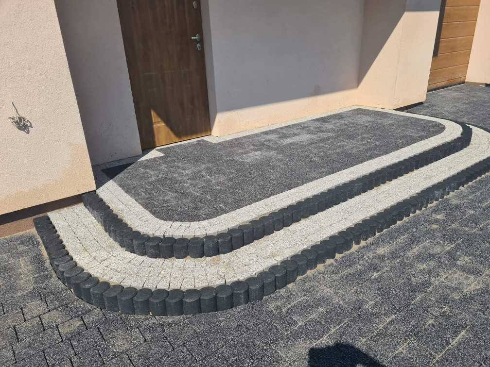 Schody wejściowe wyłożone szarą kostką brukową z obrzeżem z ciemnych, okrągłych elementów, prowadzące do drzwi wejściowych.