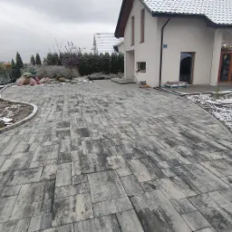 Szeroki podjazd z szarej kostki brukowej ułożonej we wzór prostokątów, obrzeża z szarej kostki, częściowo pokryty śniegiem, w tle dom jednorodzinny i rząd tuj.