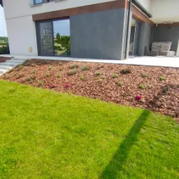 Nowoczesny ogród z trawnikiem, korą, nasadzeniami i betonowymi schodami prowadzącymi do domu z dużymi przesuwnymi oknami.