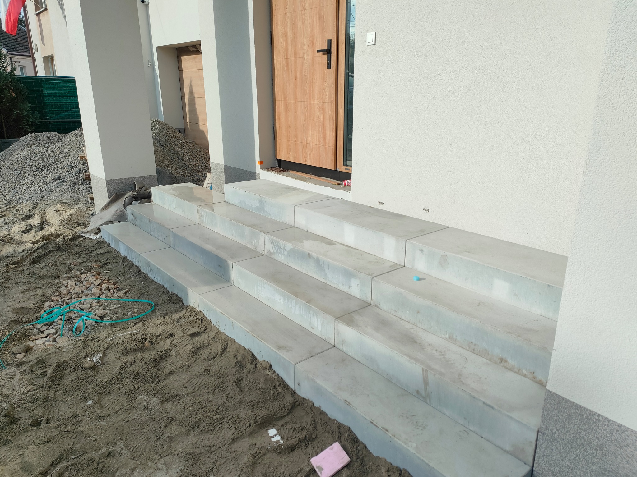 Betonowe schody wejściowe do domu w trakcie budowy, widoczny piasek i gruz, jasne wykończenie elewacji.