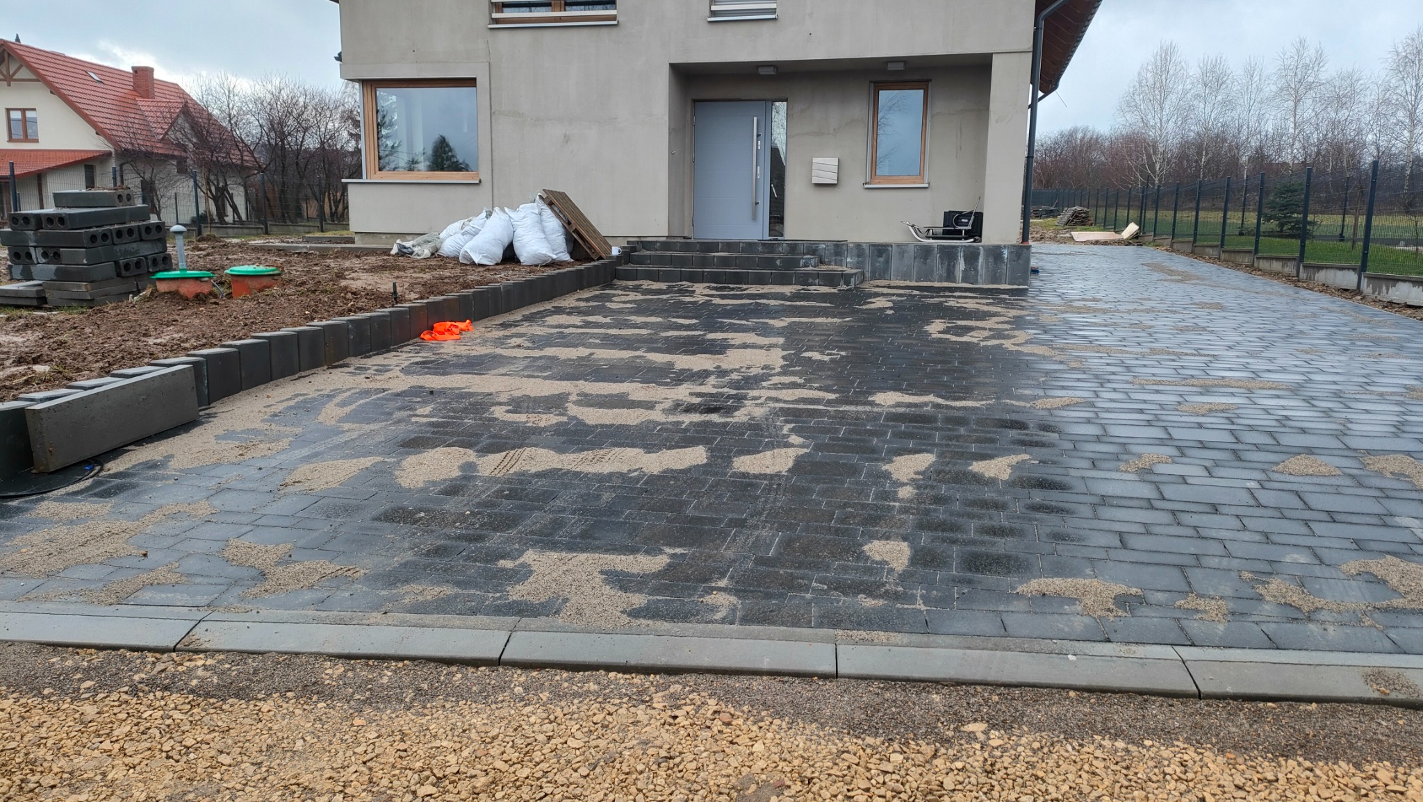Częściowo ułożona, czarna kostka brukowa na podjeździe przed domem jednorodzinnym, z widocznym piaskiem wypełniającym szczeliny i stosem kostki w tle, sugerująca trwający proces układania.