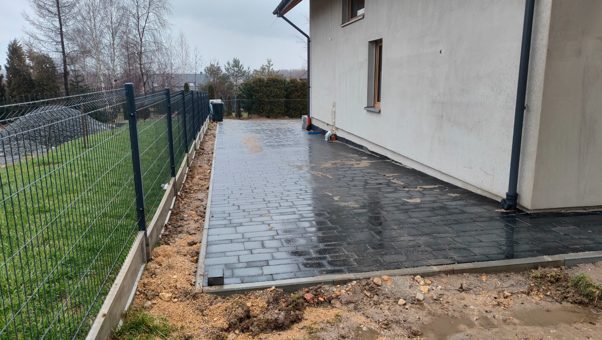 Ułożona, mokra, grafitowa kostka brukowa przy ścianie budynku i ogrodzeniu panelowym, widoczne pozostałości prac ziemnych.