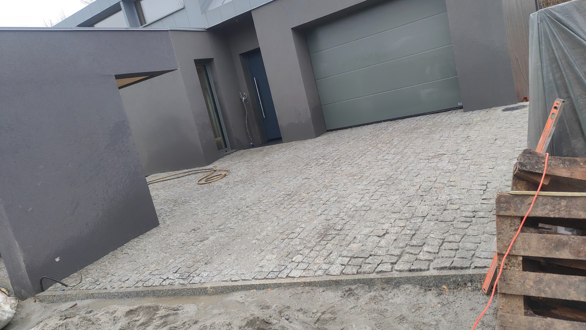 Ułożona kostka granitowa na podjeździe nowoczesnego domu w Krakowie, widoczne palety i poziomica, sugerujące trwające prace wykończeniowe.