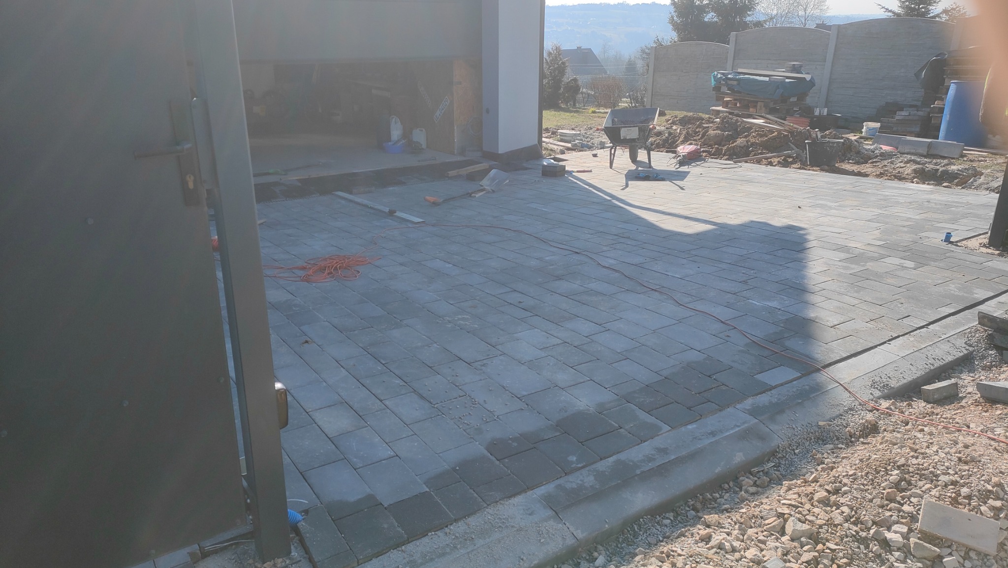 Nowo ułożona szara kostka brukowa z widocznym kablem elektrycznym, krawężnikiem i fragmentem otwartej bramy garażowej w tle, teren budowy z taczką i narzędziami.