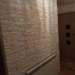 Ściana wykończona dekoracyjną okładziną imitującą kamień, z widocznym grzejnikiem i fragmentem drewnianej podłogi w tle.