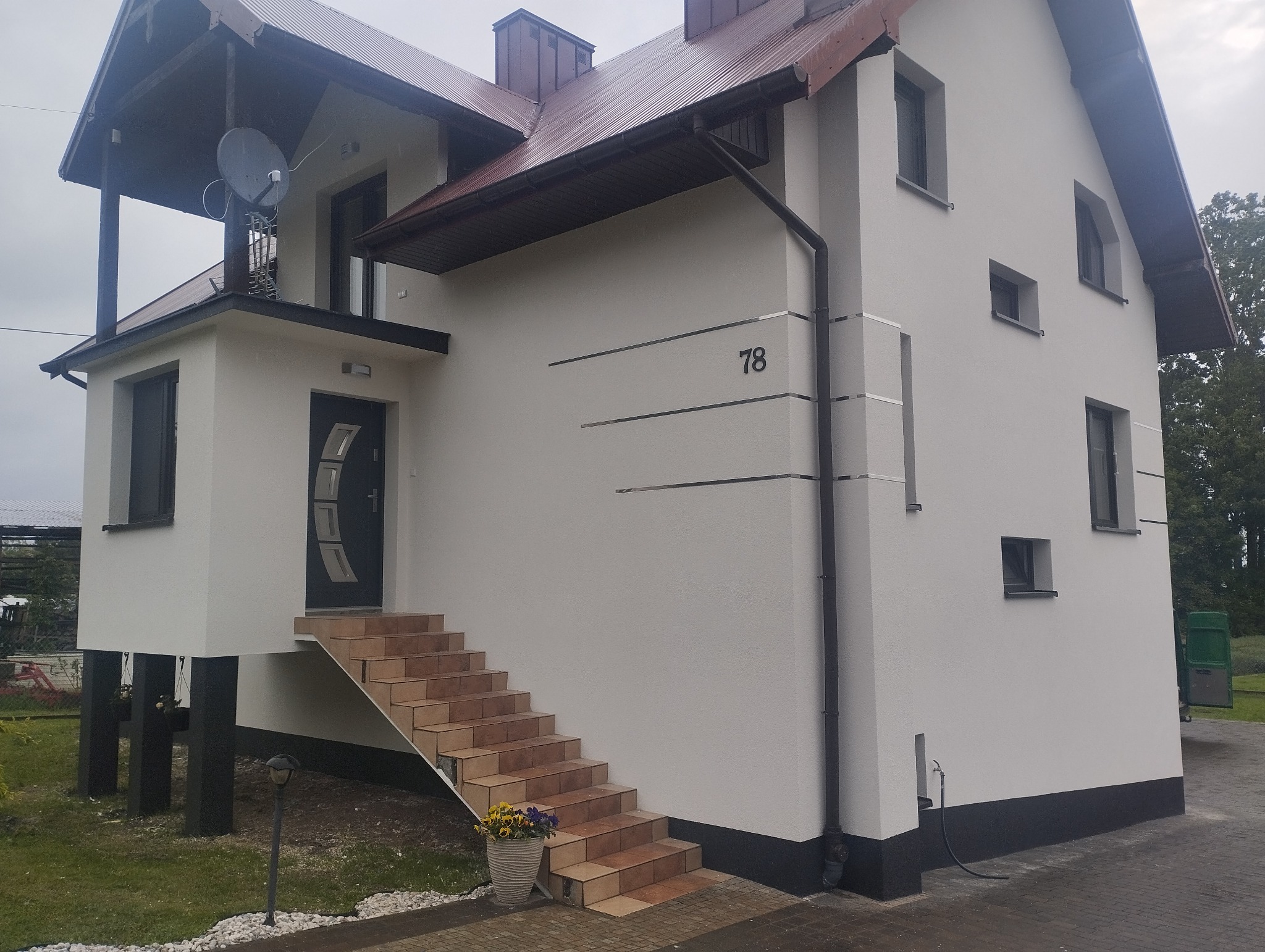 Biała elewacja domu z numerem 78 i schodami wejściowymi. Widoczne czarne rynny, brązowy dach i detale wykończeniowe. Elewacja z dekoracyjnymi, poziomymi paskami.