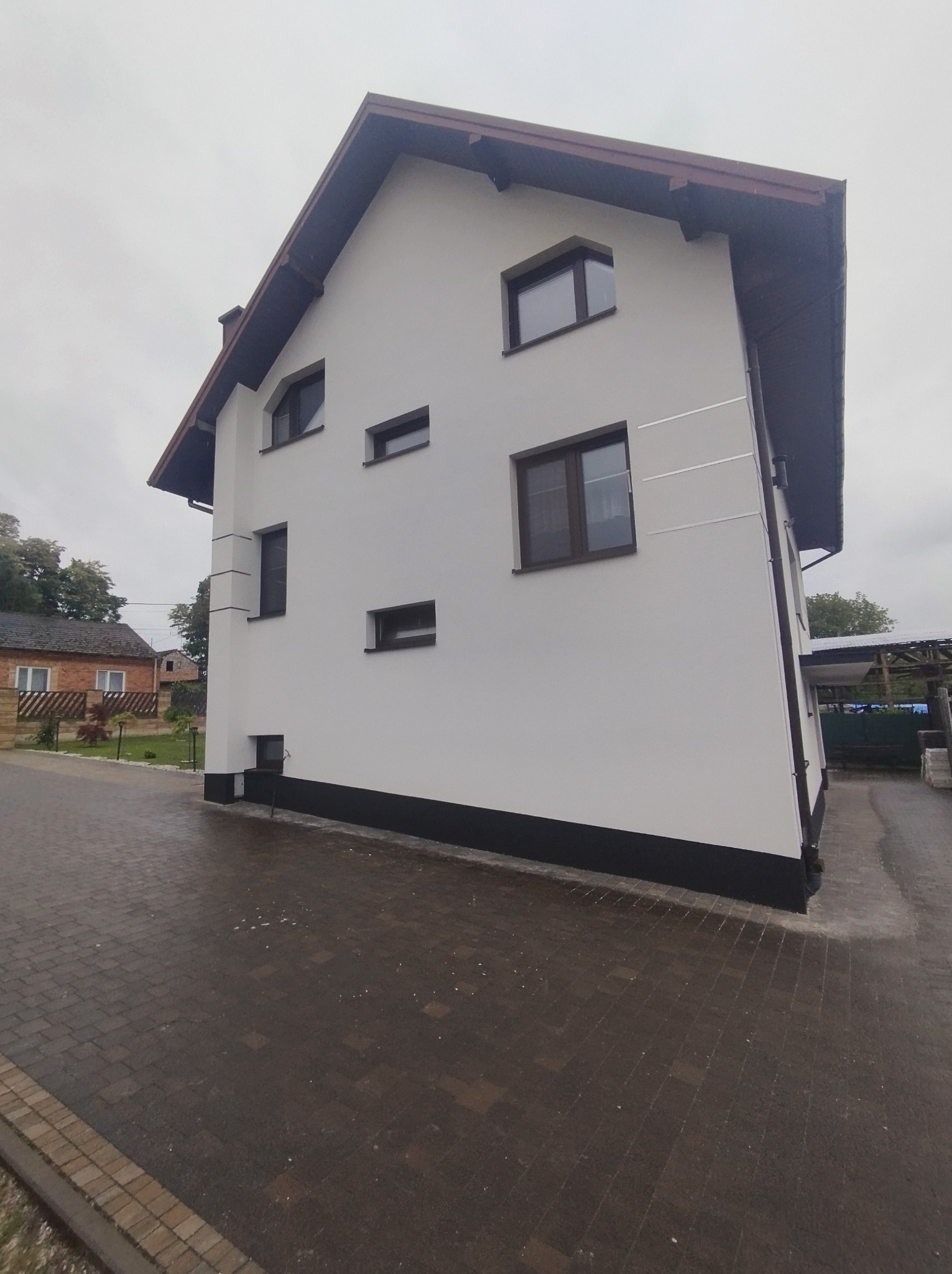 Boczny widok domu po tynkowaniu elewacji na biało z brązowymi oknami i dachem, widoczne detale wykończeniowe i kostka brukowa.