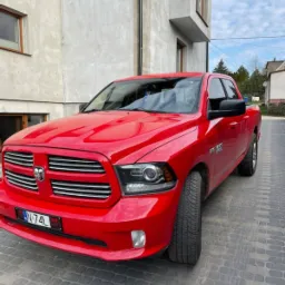 Czerwony samoch&oacute;d marki Dodge Ram zaparkowany przed nowoczesnym domem z szarego betonu architektonicznego.