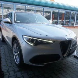 Srebrny SUV Alfa Romeo Stelvio zaparkowany przed salonem samochodowym z przeszkloną fasadą, widok z przodu, oświetlenie dzienne.